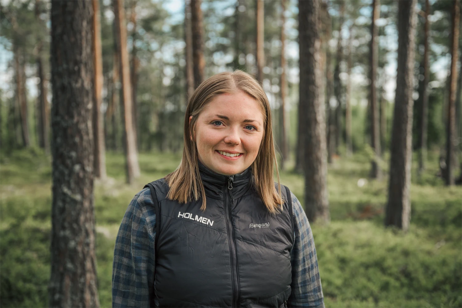 En kvinna står i skogen och tittar in kameran med ett leende.