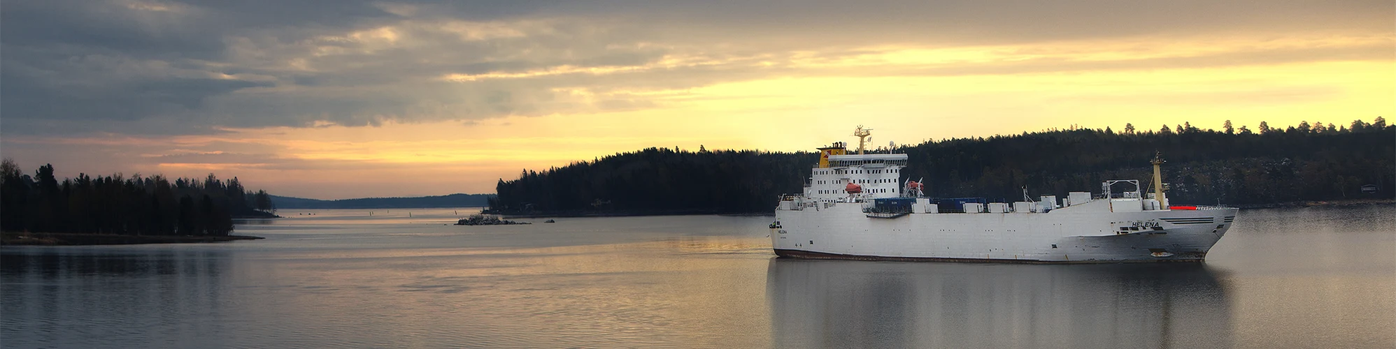 White cargo ship sailing on calm water through a coastal landscape at sunset.