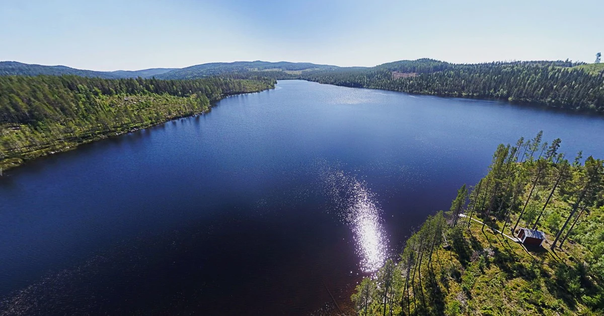 Drönarbild över kunskapsskogen Kunnådalen med en sjö i fokus. Till höger syns en röd liten stuga.